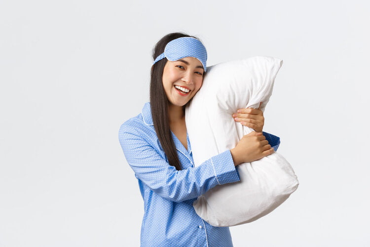 Een vrolijke jonge Aziatische vrouw in een lichtblauwe pyjama met witte stippen en bijpassend slaapmasker op haar hoofd, staat tegen een witte achtergrond en omhelst een wit kussen. Ze lacht breed en lijkt zich op een comfortabele en ontspannen slaap te verheugen.