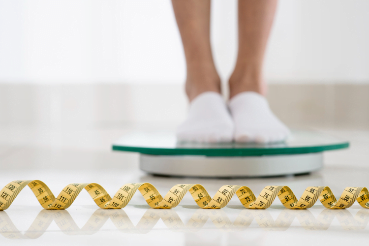 Close-up view of a person's lower legs and feet standing on a bathroom scale, with white slippers on, above a coiled yellow measuring tape on a white tiled floor, indicating a concept of weight measurement and management.