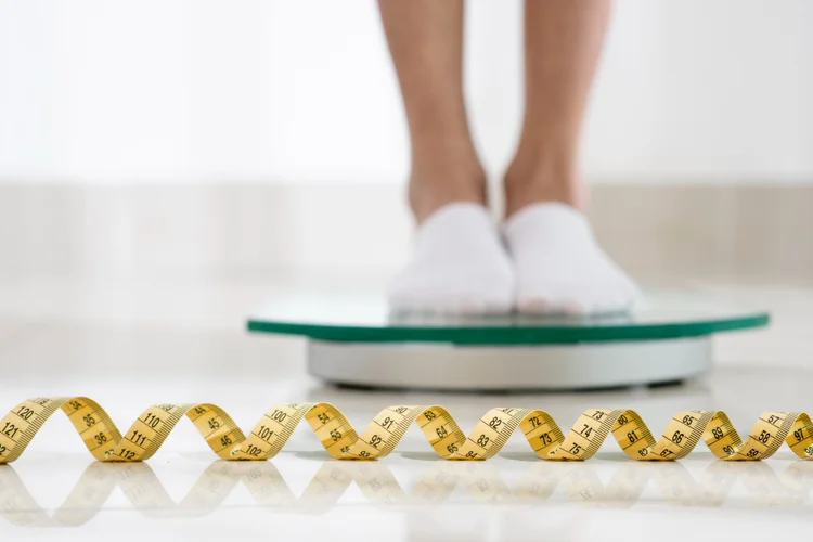 Close-up view of a person's lower legs and feet standing on a bathroom scale, with white slippers on, above a coiled yellow measuring tape on a white tiled floor, indicating a concept of weight measurement and management.