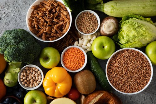 Assortiment van gezonde voedingsmiddelen waaronder groente, fruit, peulvruchten en volkoren pasta op een grijze ondergrond.
